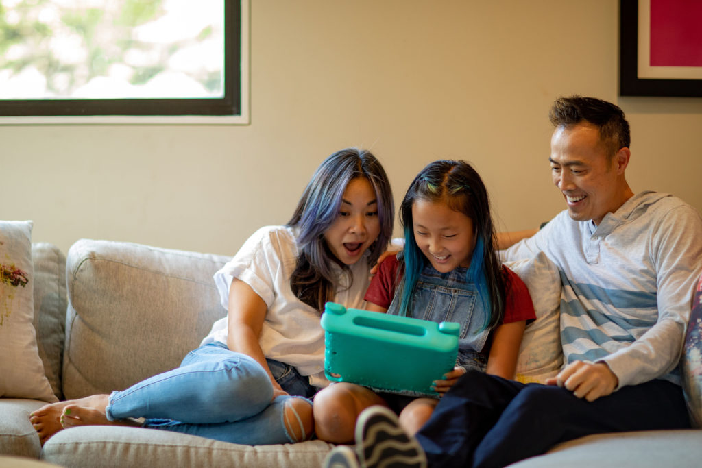 Family sitting and smiling together on their cream-colored couch. The pre-teen girl is showing her parents the video games she’s playing on Roblox with her tablet. Mom has a surprised look as she realizes how much her daughter is learning from video games.