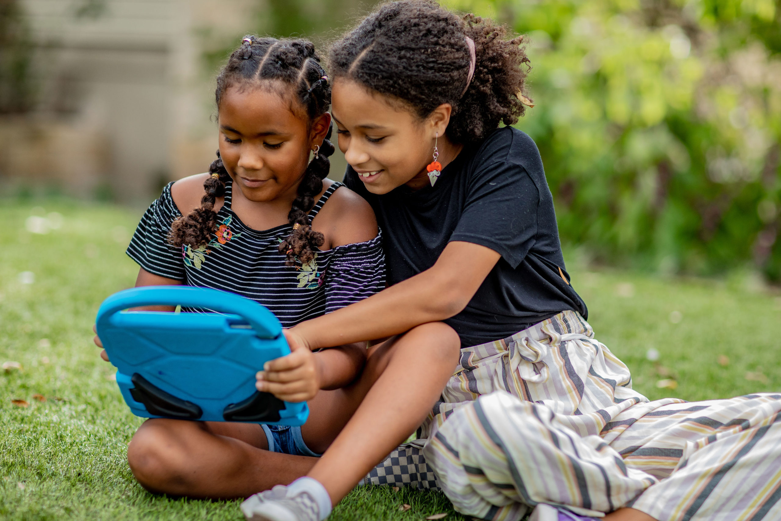 Two young girls sit outside playing video games on a blue tablet. This is Learnified Gaming! One girl is excitedly pointing to the screen as they play together. Video games are a fun way to build social skills, both online and in person.