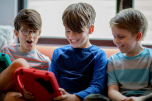 Three young boys playing video games together on their tablets, smiling and excited. They’re practicing and improving their psychomotor speed and other life skills while they play together. One looks super impressed at what the other is doing in his video game.