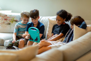 Four kids playing Roblox together on a beige couch while socializing. Play-based learning and social interaction can look different than wooden toys or kids on a playground.