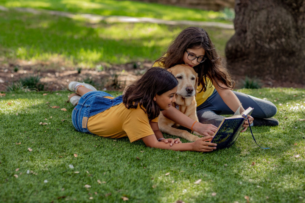 Two girls and a golden retriever sitting in the grass and reading a book. There are lots of ways to have fun with reading!