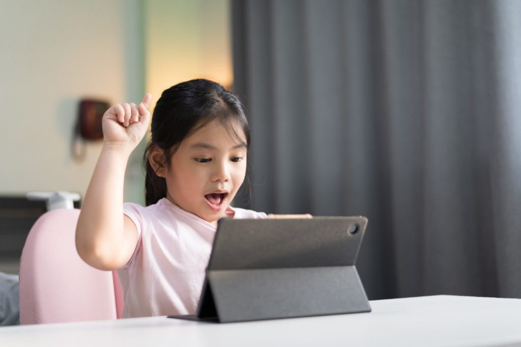 A young girl is sitting at a table and playing on her tablet. She looks excited with a fist raised in the air in triumph. She's practicing goal setting in her video game, and just achieved a big win!