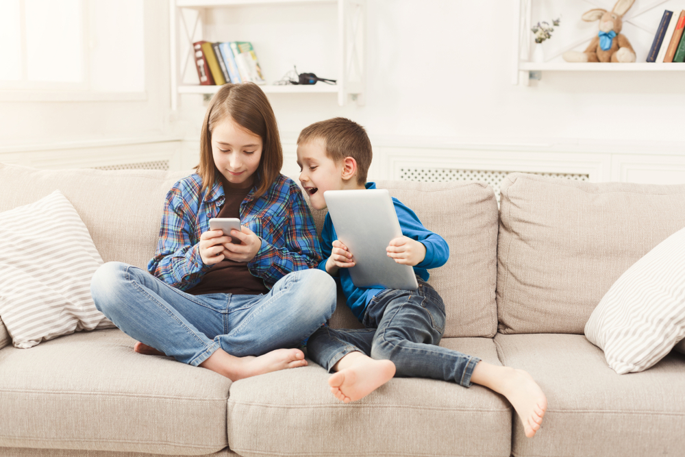 Two kids, older sister and her brother, having screen time together on a beige couch. The boy is smiling big while looking at his sister's phone.