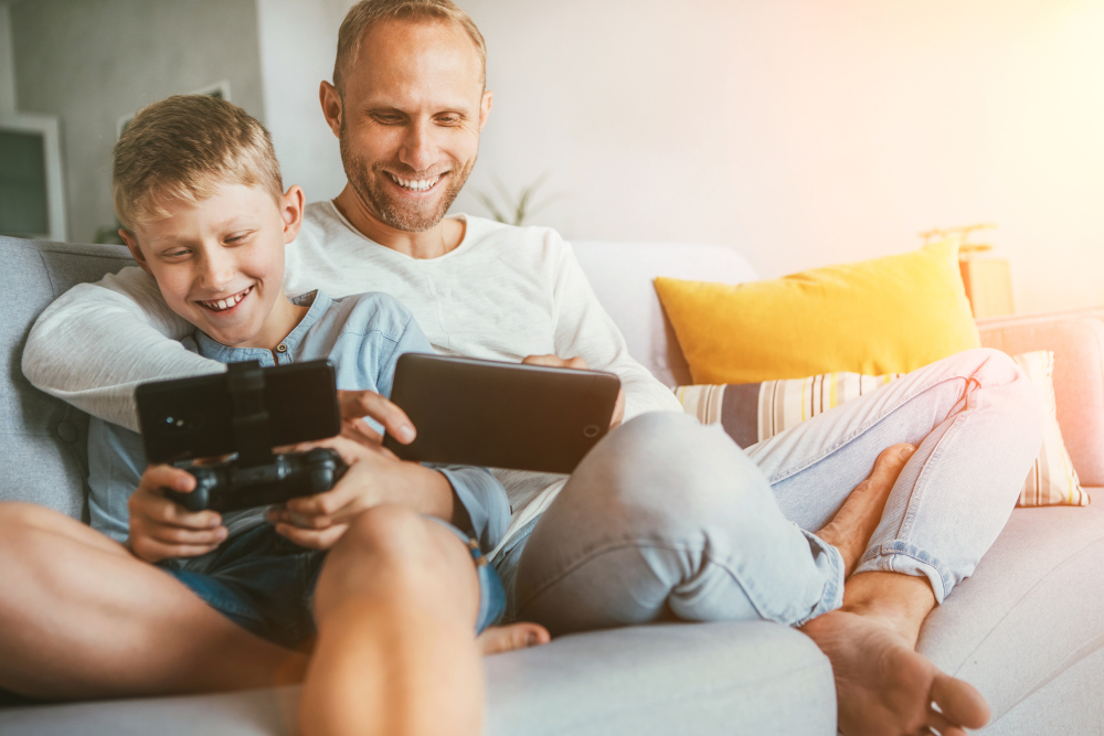 Dad and his kid are sitting on a couch together, smiling while playing on their devices. The kid may be happy because Dad is adding Robux to his Roblox account.