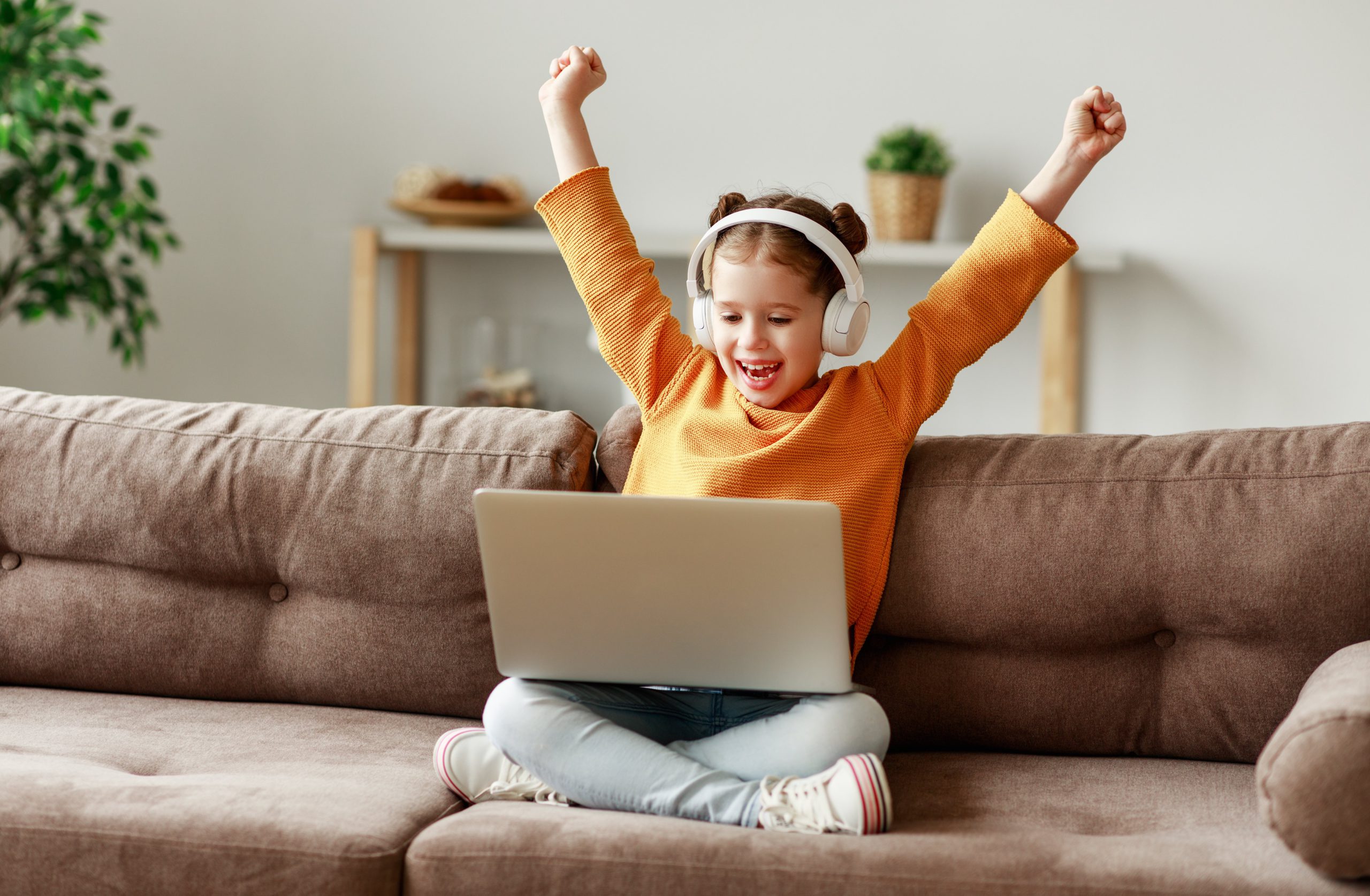 Happy girl in headphones raising arms and smiling while sitting crossed legged on sofa and celebrating victory in video game on laptop. Using tech for learning can help keep kids engaged and inspired.