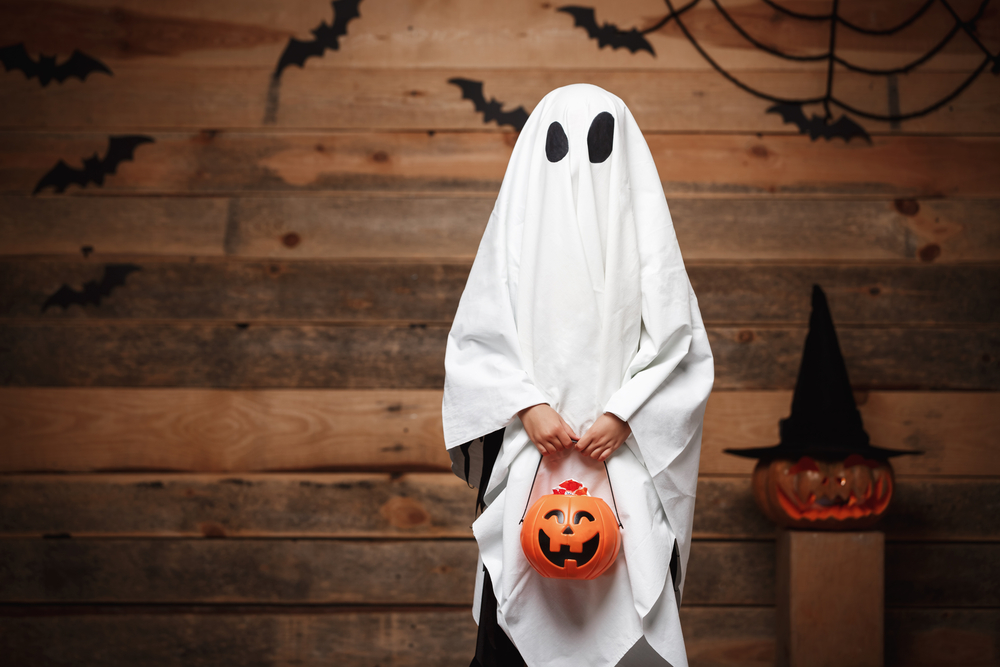 Image of a child in a ghost halloween costume holding a bucket to collect candy. This kid loves Halloween and spooky Roblox games!
