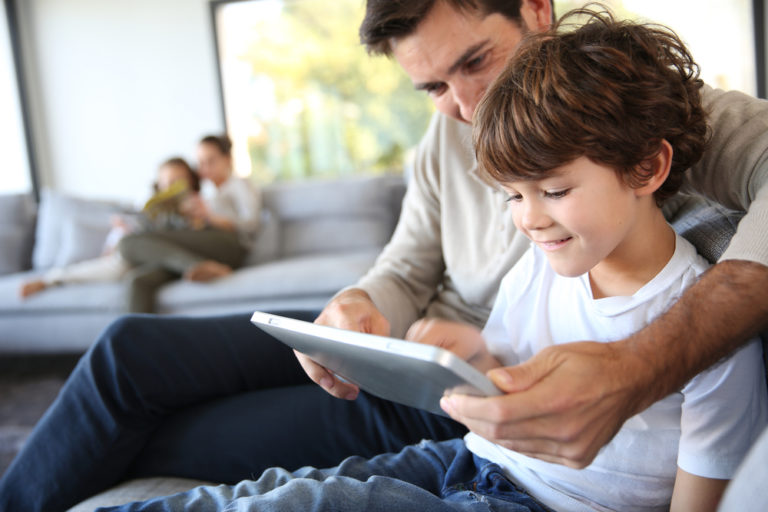 A father and son sit on the couch playing on a tablet.
